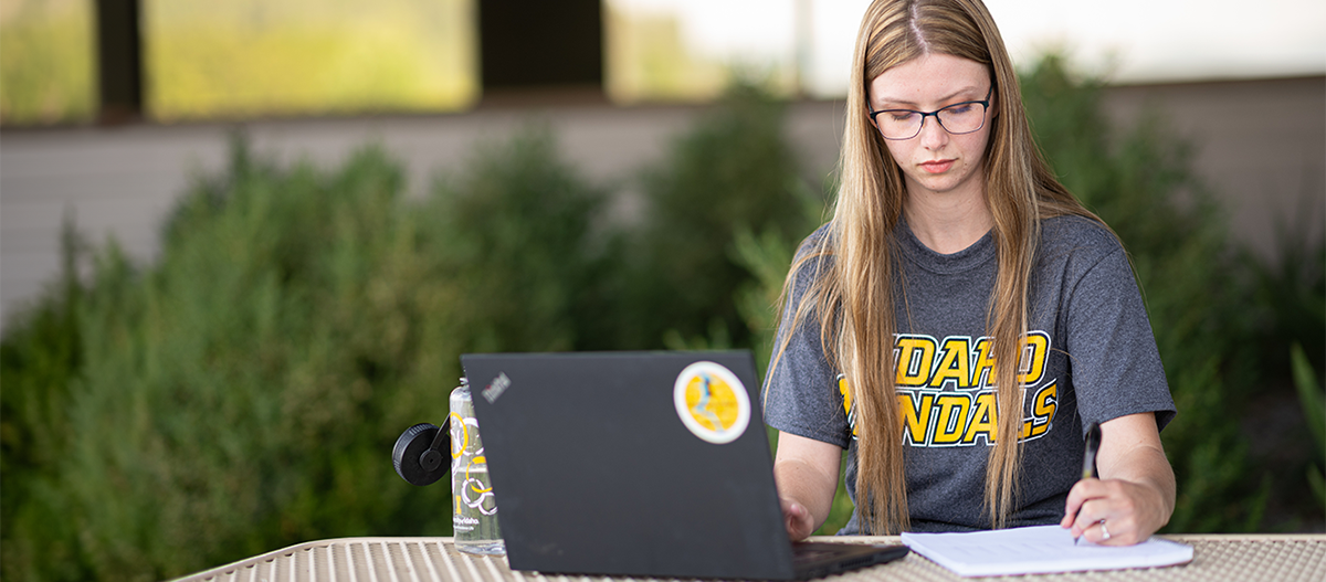 Girl writing notes from her laptop outside on a table.