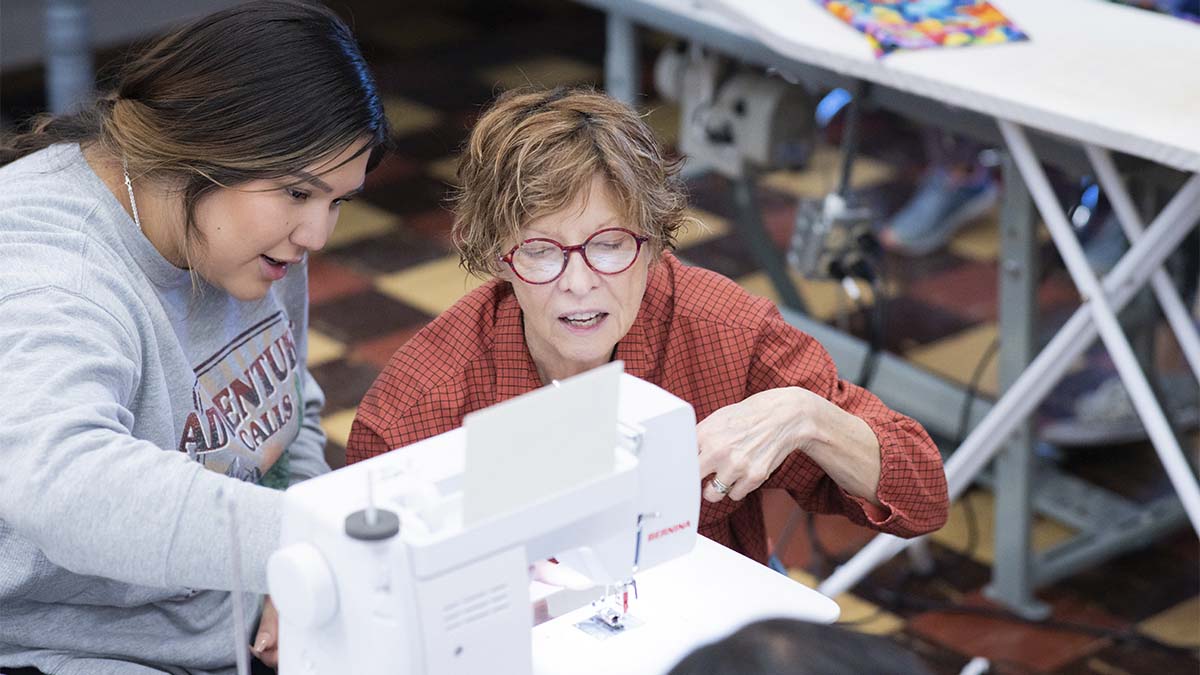 A woman teaches a student how to use a sewing machine.