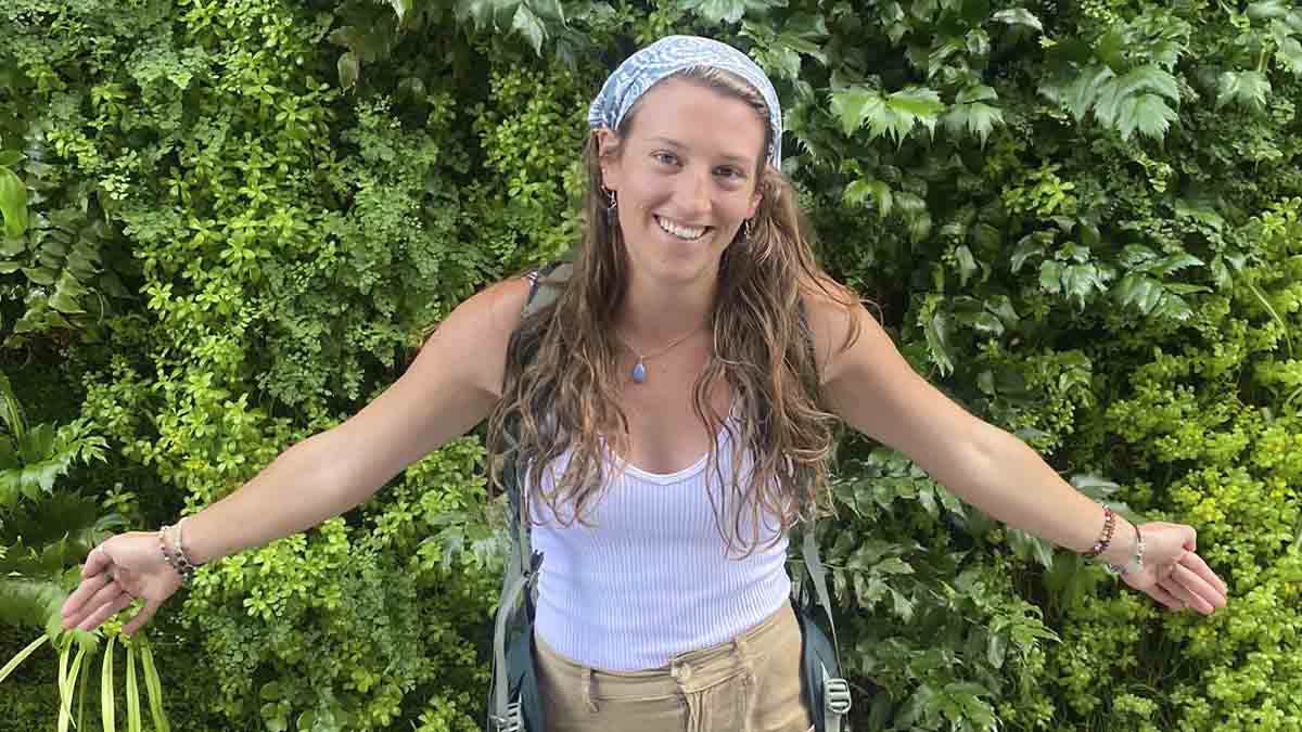 A woman smiles as she poses in front of a wall of green plants.