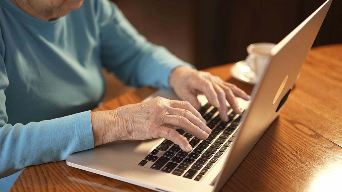 An elderly woman’s hands rest on the keyboard of a laptop.