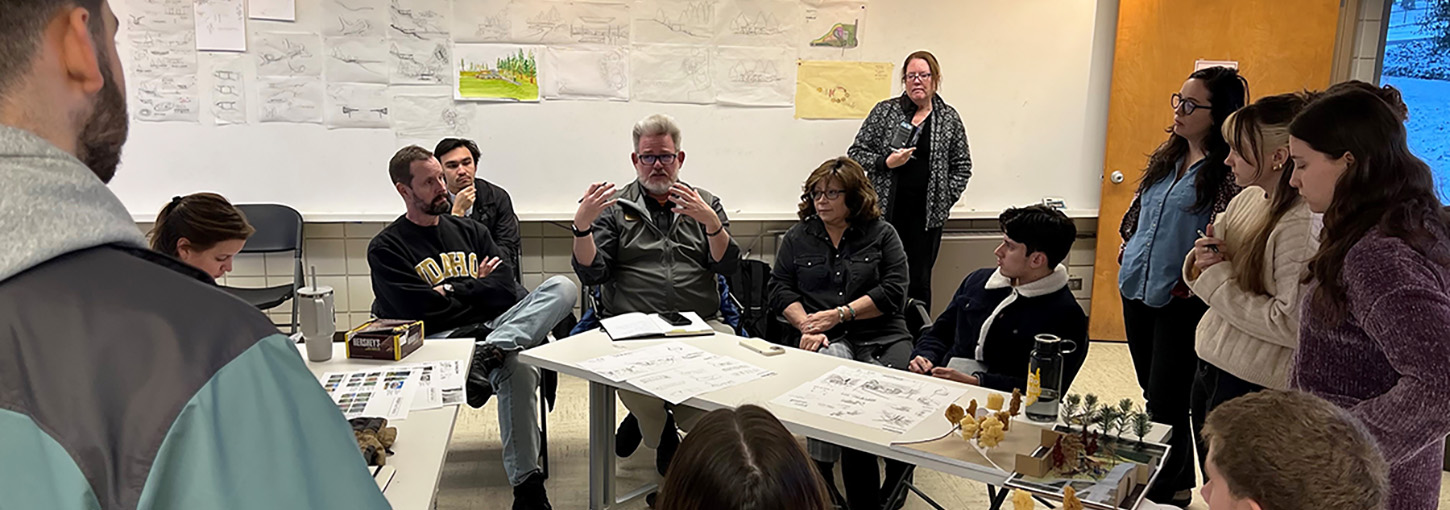 Photo of students and faculty sitting around tables with drawings in the background.