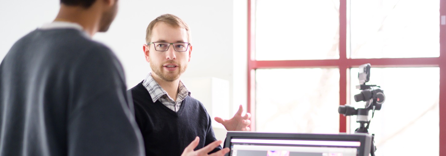 Researcher Damon Woods smiles and looks into the camera in a brightly lit, yellow and beige striped room with large windows.